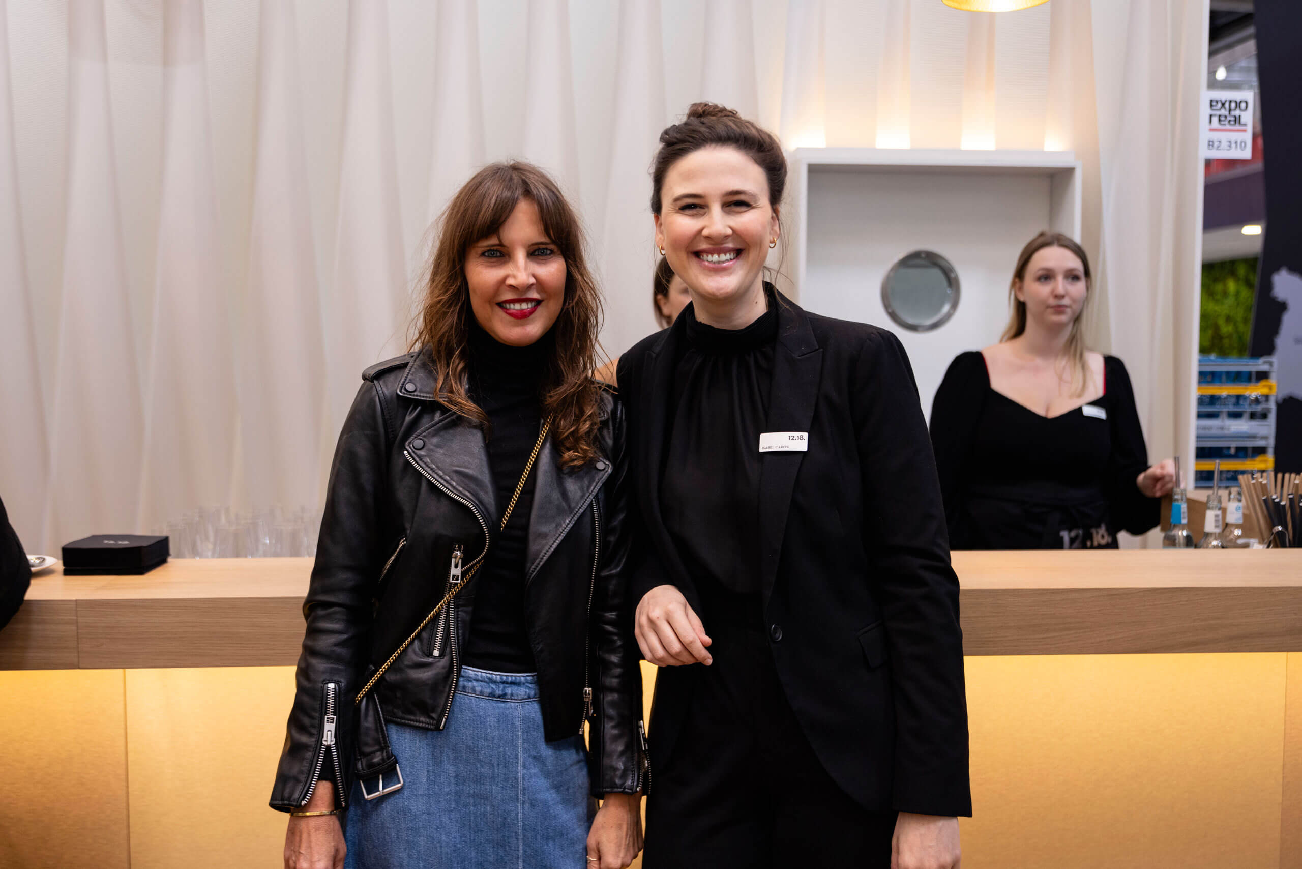 Zwei lächelnde Frauen stehen zusammen in einem Innenraum, EXPO im Hintergrund