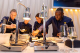 Eine Gruppe von Personen in Schürzen beim Kochen in einer Küche mit Tischgeschirr und Flaschen